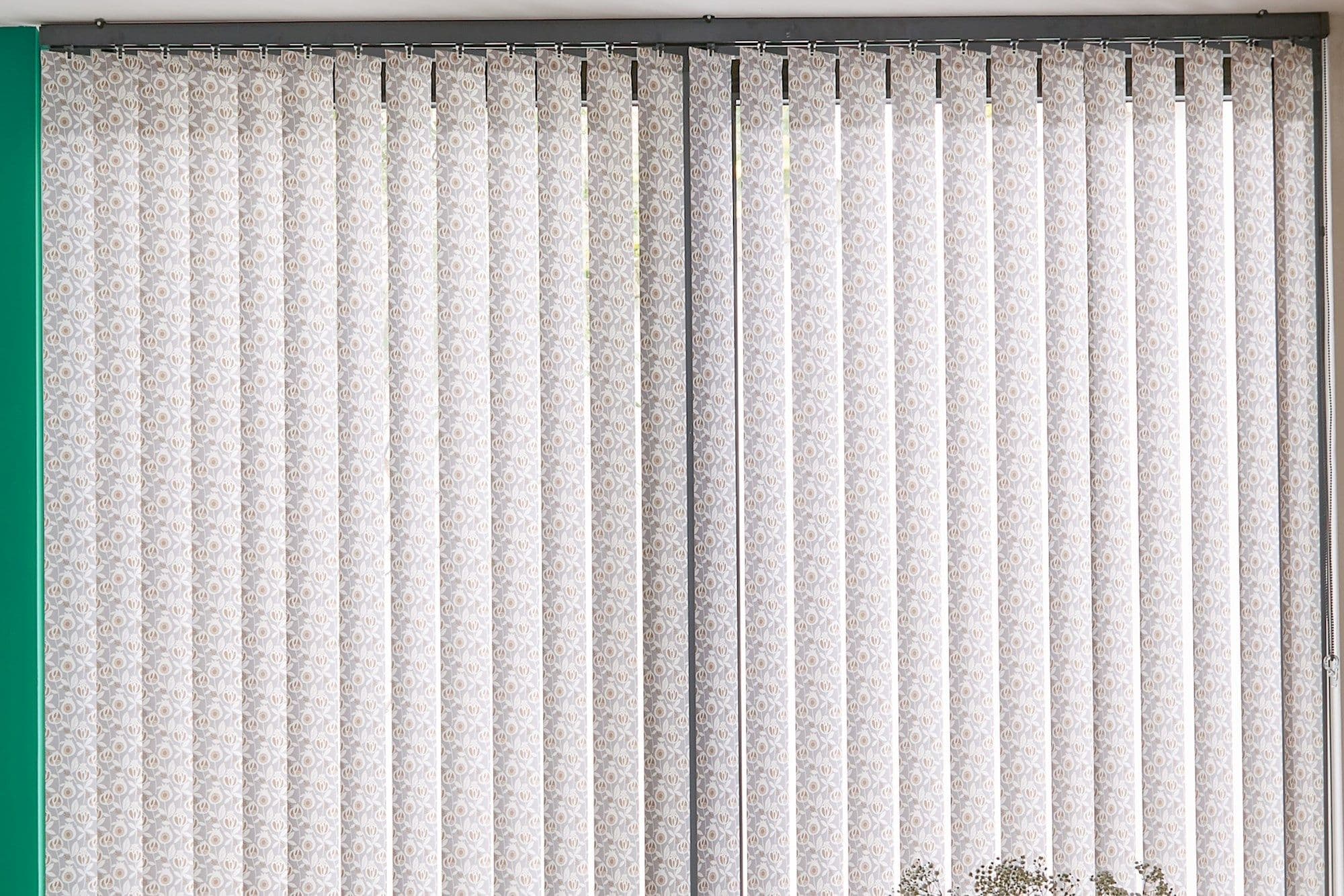 Vertical fabric blinds covering wide window; patterned, closed slats filtering daylight; mounted on top rail with side chain; interior with green left wall and small potted plant.