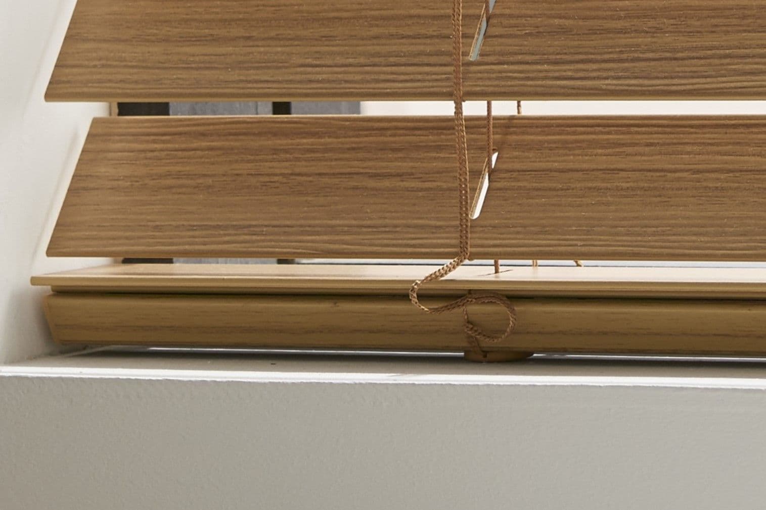 Wooden horizontal slatted blinds, lowered and slightly tilted with cords looped and hanging; blocking most light through a white window frame, interior wall and windowsill visible.