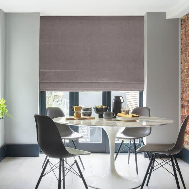 A taupe fabric Roman blind, fully lowered with horizontal folds, filtering daylight over glass French doors; dining area with white tulip table, four gray chairs, exposed brick and patio beyond.