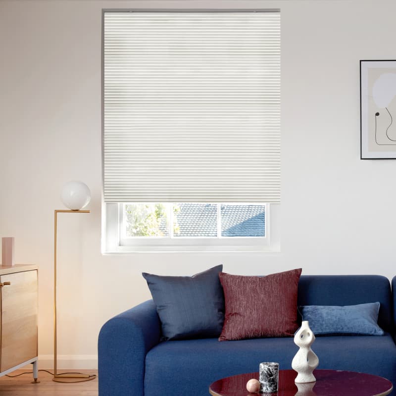 Off-white pleated cellular blind, lowered across the window with a small bottom gap, filtering daylight; positioned above a navy sofa with throw pillows, floor lamp, sideboard, and minimal living-room decor.