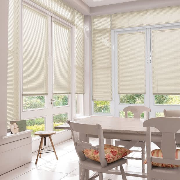 Object â€” Cream pleated blinds on tall windows and French doors. Action â€” Mostly lowered, filtering daylight through small gaps. Context â€” Bright dining nook with white table, chairs, floral cushions, garden view.