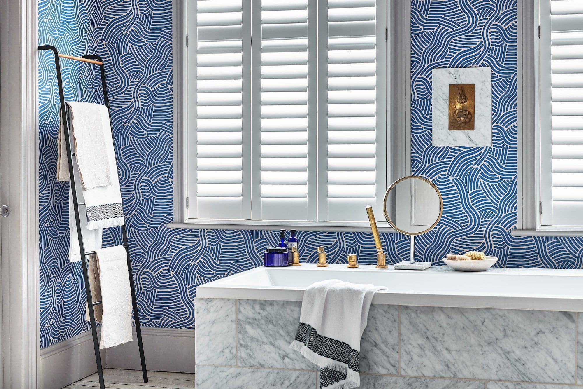 White plantation shutters covering the central window, wide horizontal louvers closed and softly diffusing daylight; bathroom interior with a marble-faced bathtub, brass fixtures, blue patterned wallpaper, and towel ladder.