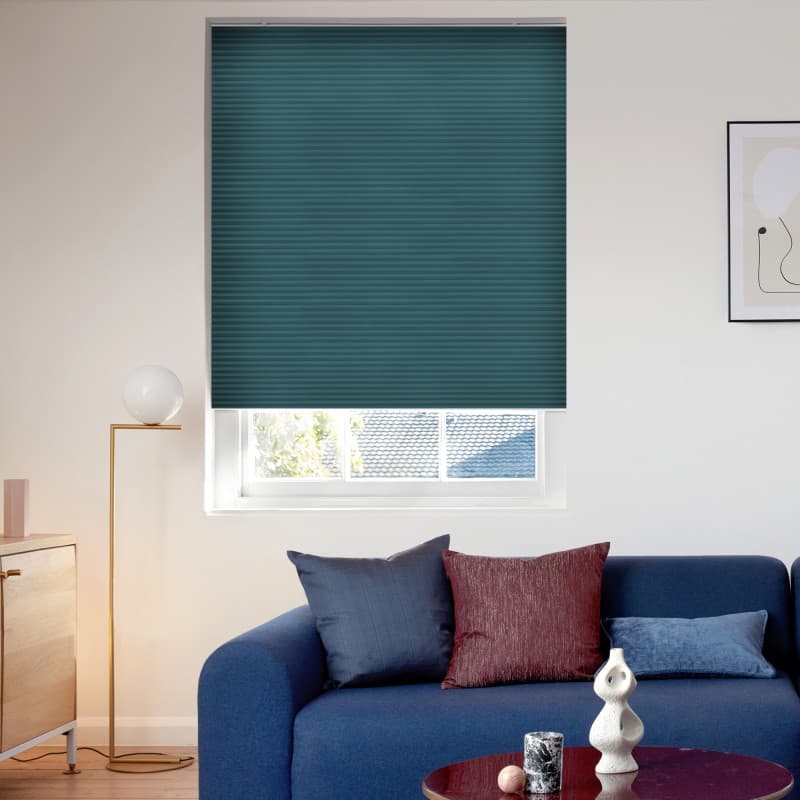 Teal pleated cellular blind lowered over a tall window, fully closed and dimming light; in a sunlit living room with a blue sofa, cushions, floor lamp, and framed abstract art.