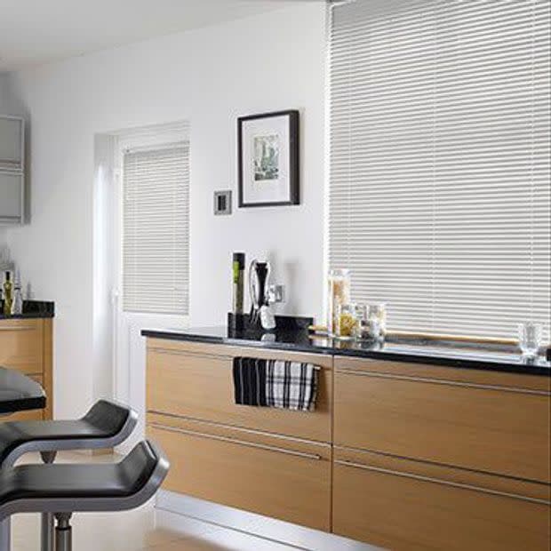 White horizontal blinds lowered over a large window and adjacent door, closed and softly filtering daylight; modern kitchen with light wood cabinets, black countertop, two bar stools and minimal decor.