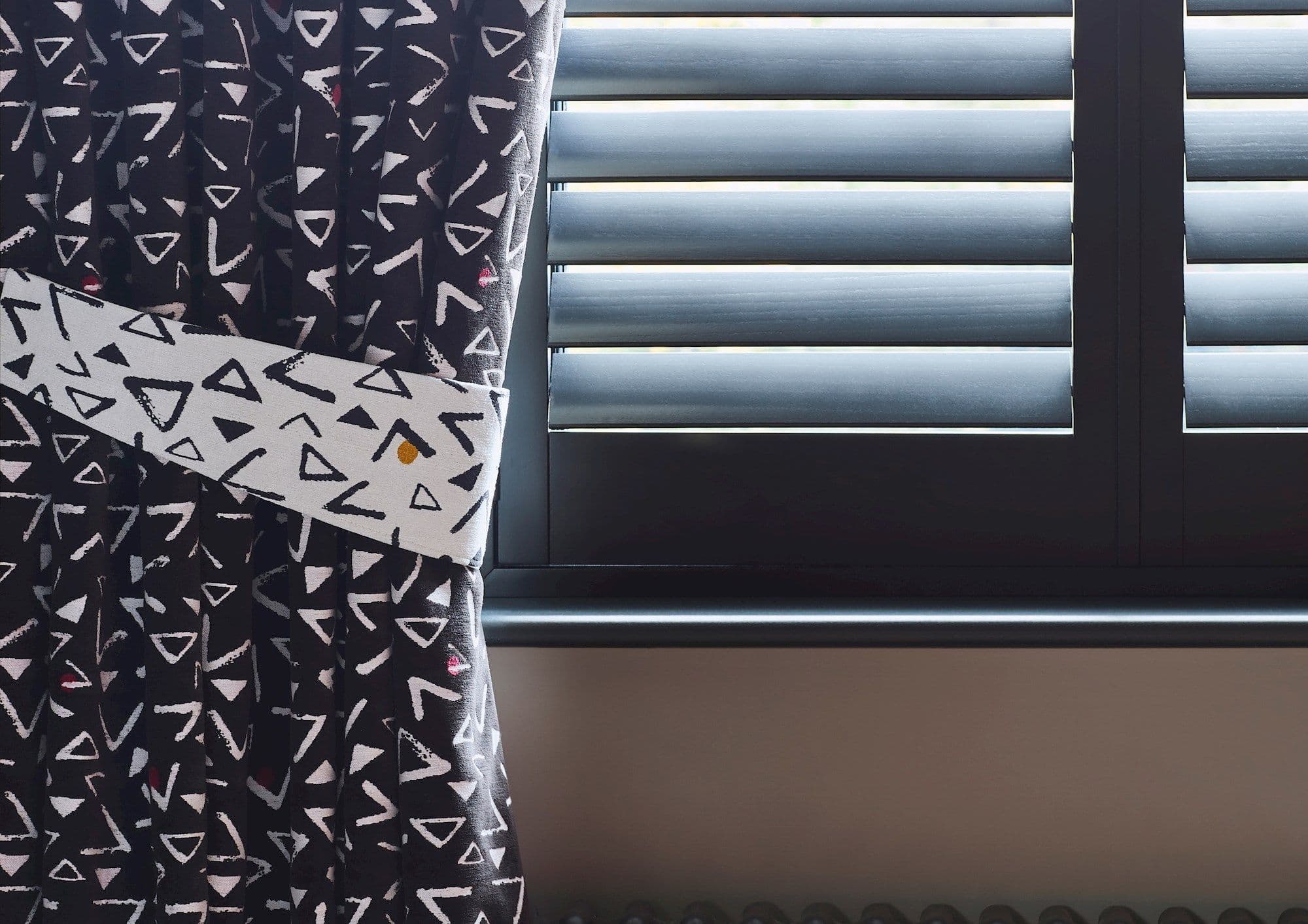 Black curtain with white triangular print, held back by a matching tieback; pulled aside to reveal closed horizontal shutters whose angled slats filter daylight; interior, windowsill visible.