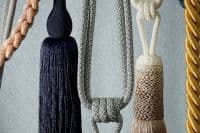 Decorative curtain tassels and braided tiebacks, assorted colors (navy, beige, gold), hanging and looped with knots and braids in front; close-up against a pale textured wall under soft light.
