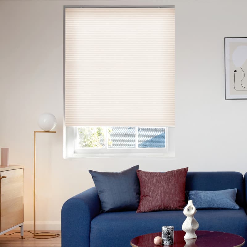 Light beige pleated honeycomb blind lowered over the window, softly filtering daylight; modern living room with navy sofa and cushions, brass floor lamp, wooden sideboard and framed wall art.
