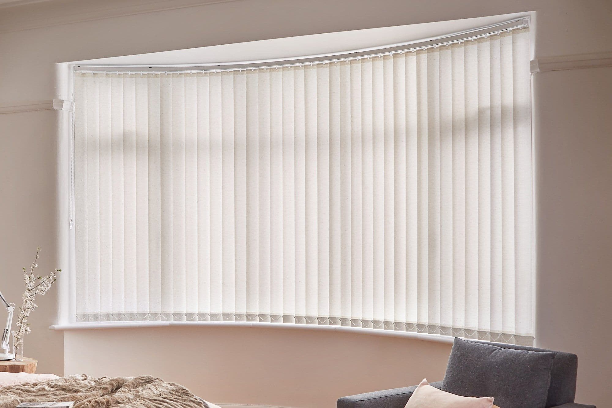 Vertical fabric blinds across a curved bay window, fully closed and diffusing soft daylight; set in a softly lit bedroom corner with a bed, bedside lamp and upholstered chair.
