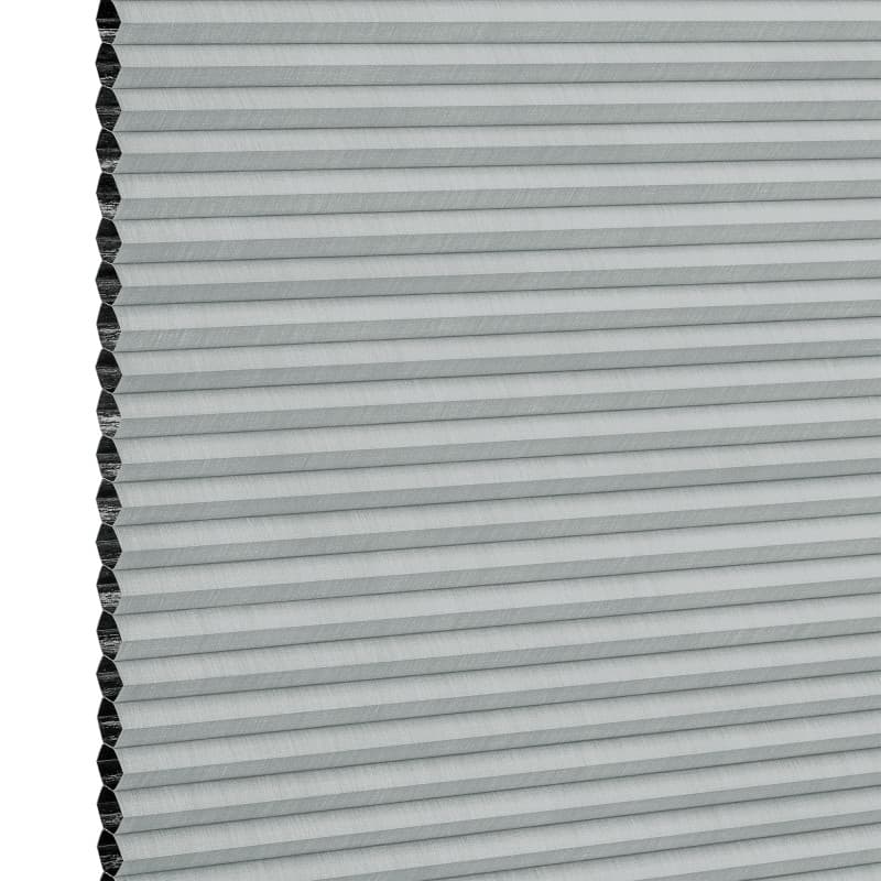 Light-gray honeycomb (cellular) blind, mounted vertically with narrow horizontal pleats and exposed hexagonal cells at the left edge; fully closed, filtering light; close-up view against a white background.