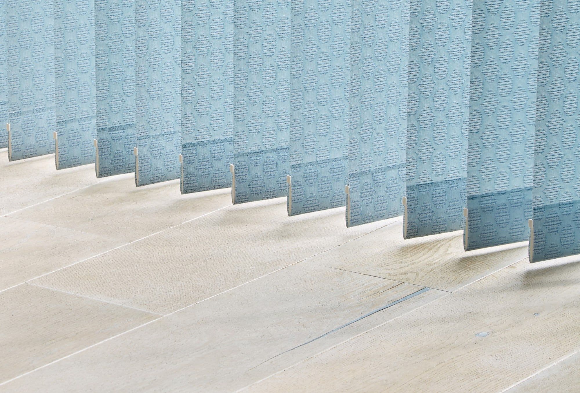Light-blue patterned vertical blinds, hanging in a row with visible weighted bottoms; partially closed, filtering soft daylight onto a pale wooden floor in a bright interior.