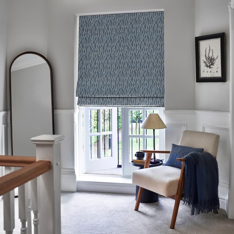 Patterned blue Roman blind lowered over French doors, folding near the bottom; filtering daylight into a carpeted landing nook with an armchair, side table, lamp, mirror and artwork.