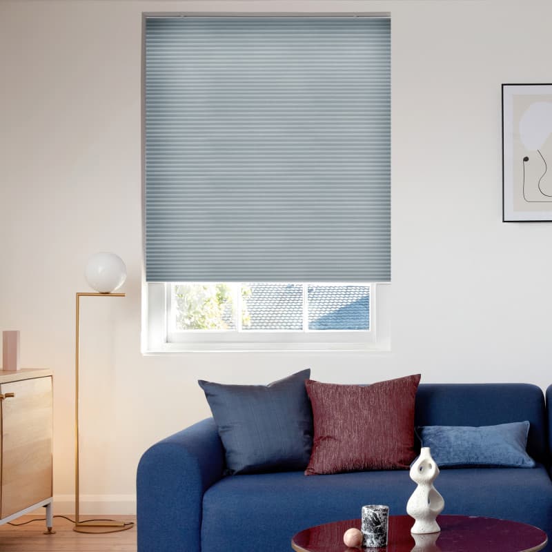 Light-gray pleated cellular blind lowered over the window, filtering daylight through horizontal ribs; above a blue sofa in a modern living room with cushions, floor lamp, and partial roof-and-tree view.