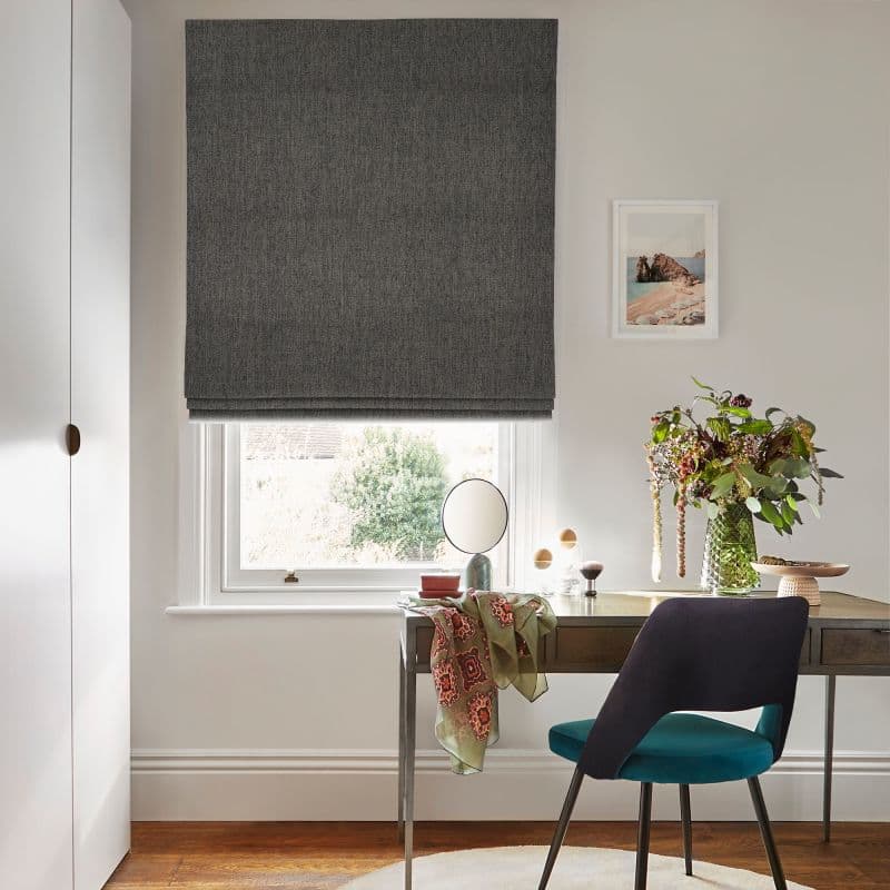Dark grey Roman shade, fully lowered with subtle folds at the bottom, filtering daylight through a white sash window; above a wooden desk with teal chair, vase of flowers and a small mirror.