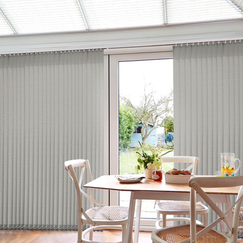 Grey fabric vertical blinds, floor-to-ceiling with slim slats and visible headrail; mostly closed but parted centrally, filtering daylight into a bright dining nook with wooden table, chairs, and a garden view.