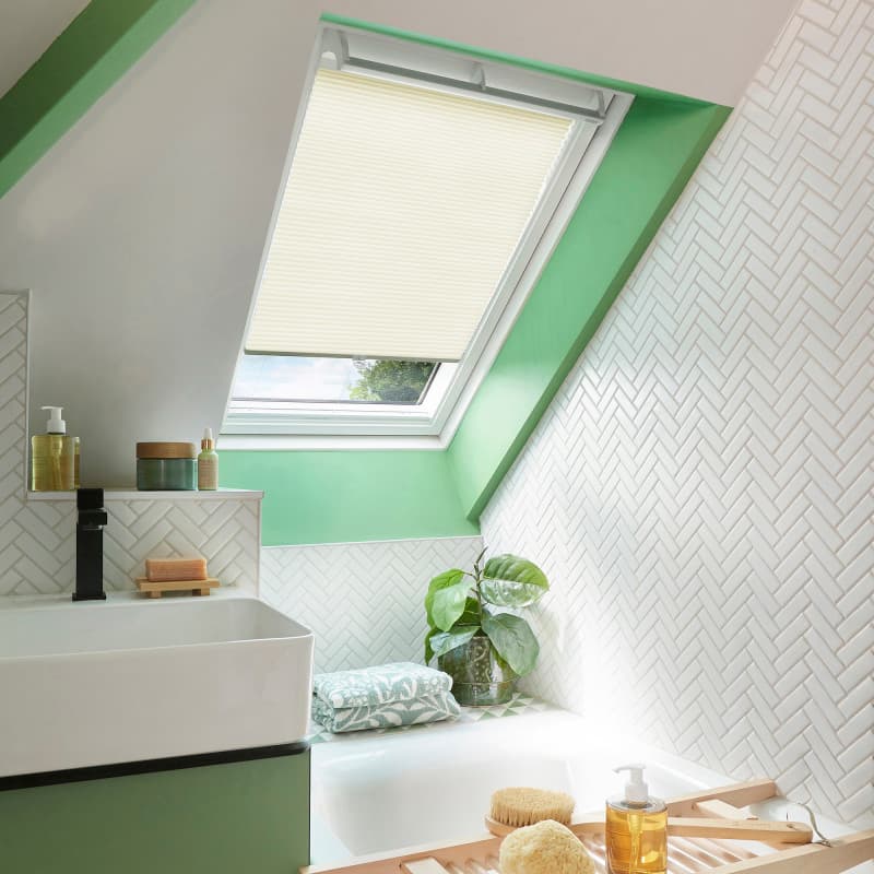 Cream pleated blind on a slanted skylight, partially lowered and filtering daylight. Sloped attic bathroom with green window trim, white herringbone-tiled walls, sink, bathtub, plant and bath tray.