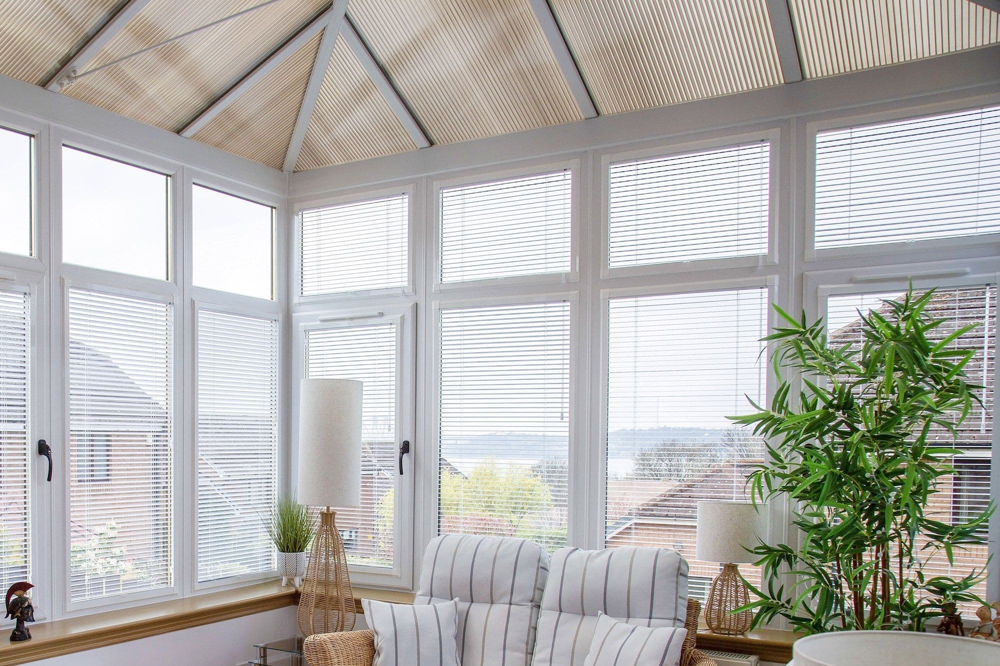 White horizontal blinds cover tall multiâ€‘pane conservatory windows, closed and filtering bright daylight; room contains striped cushioned rattan chairs, wicker lamps, a potted bamboo and suburban roof views.