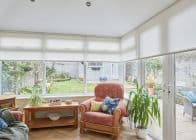 Cream roller blinds cover the upper halves of corner conservatory windows, lowered and softly filtering daylight; inside, an upholstered armchair, wooden table, potted plants, and a small garden beyond.