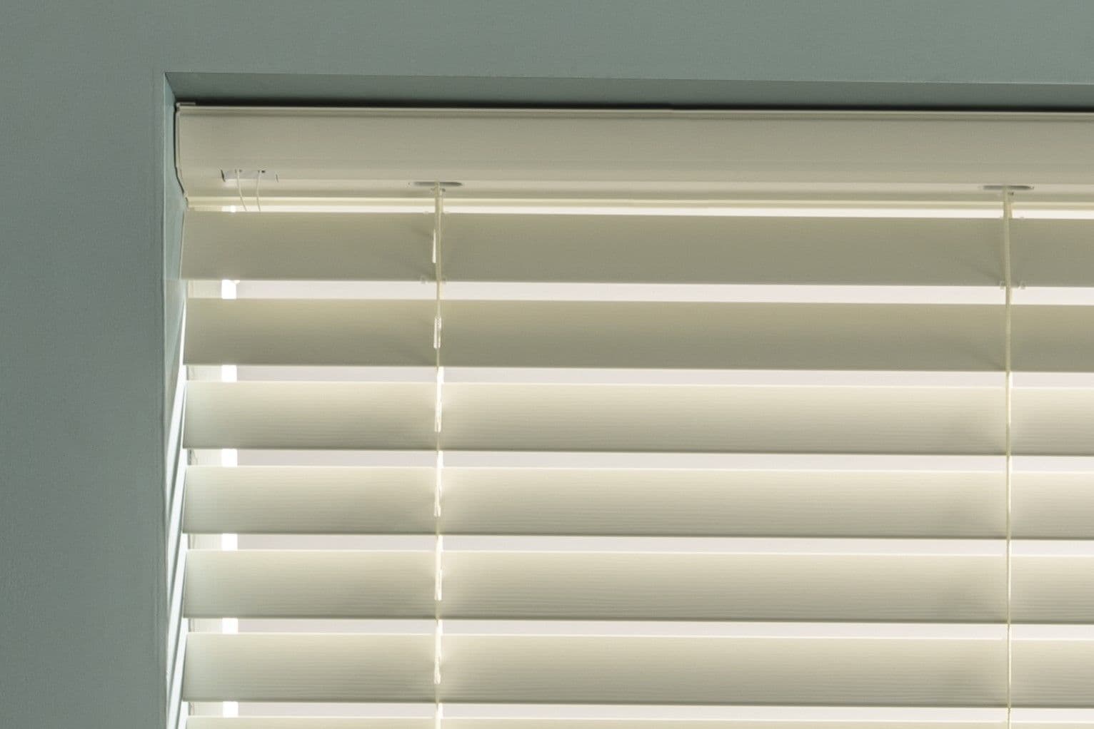 White horizontal slatted blinds occupying the top of a recessed window, partially closed with visible ladder tapes and pull cords, filtering bright daylight in a pale, softly lit room.
