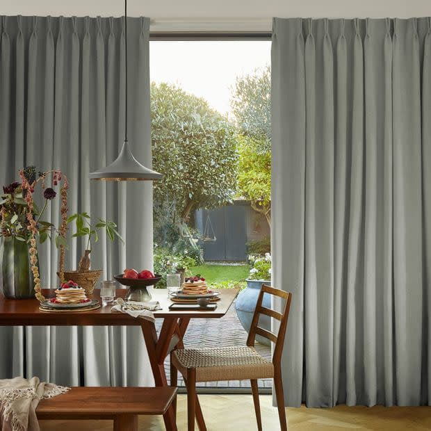 Floor-length light-gray pinch-pleat curtains, mostly drawn aside in a centered gap, softly filtering daylight and framing a doorway; dining table and chairs set with breakfast in a sunlit garden-facing room.
