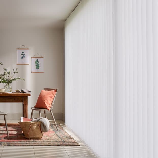 White vertical blinds covering a floor-to-ceiling window, closed and softly filtering sunlight; dining nook with wooden table, chair with terracotta cushion, patterned rug, wicker basket, botanical prints, bright daylight.