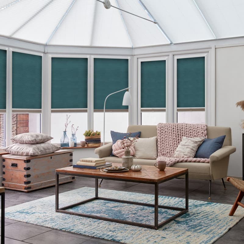 Teal pleated cellular blinds, fitted inside white conservatory window frames and lowered, filtering daylight; in a cozy conservatory living area with a beige sofa, pink knit throw, and wooden coffee table.