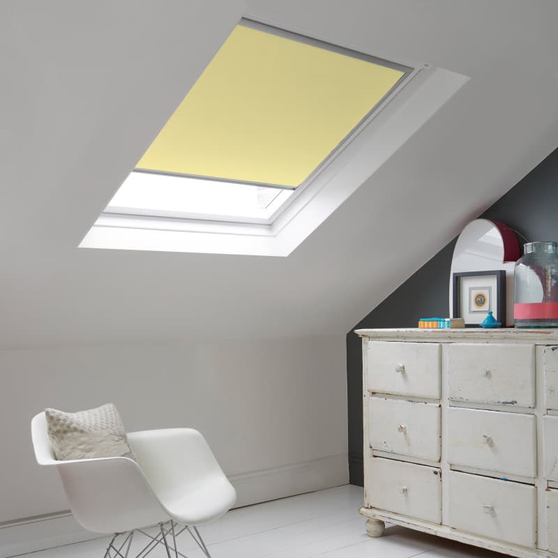 Room with white eaves wall, dark blue-grey wall to right with a shabby chic white drawer unit, a contemporary white chair, white painted floorboards and a skylight blind in lemon.