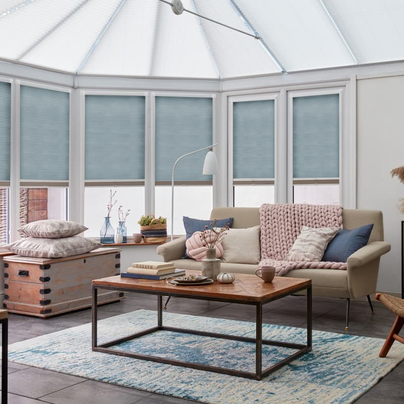 Pale blue cellular blinds covering conservatory windows, lowered and gently filtering daylight; above a cozy sunroom with a beige sofa, pink chunky knit throw, wooden coffee table and blue rug.