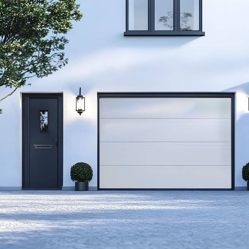 White sectional garage door (right of a dark entry door) â€” closed and flush within dark trim; modern white facade with wall lanterns, potted shrubs, and a tree casting soft daylight shadows.
