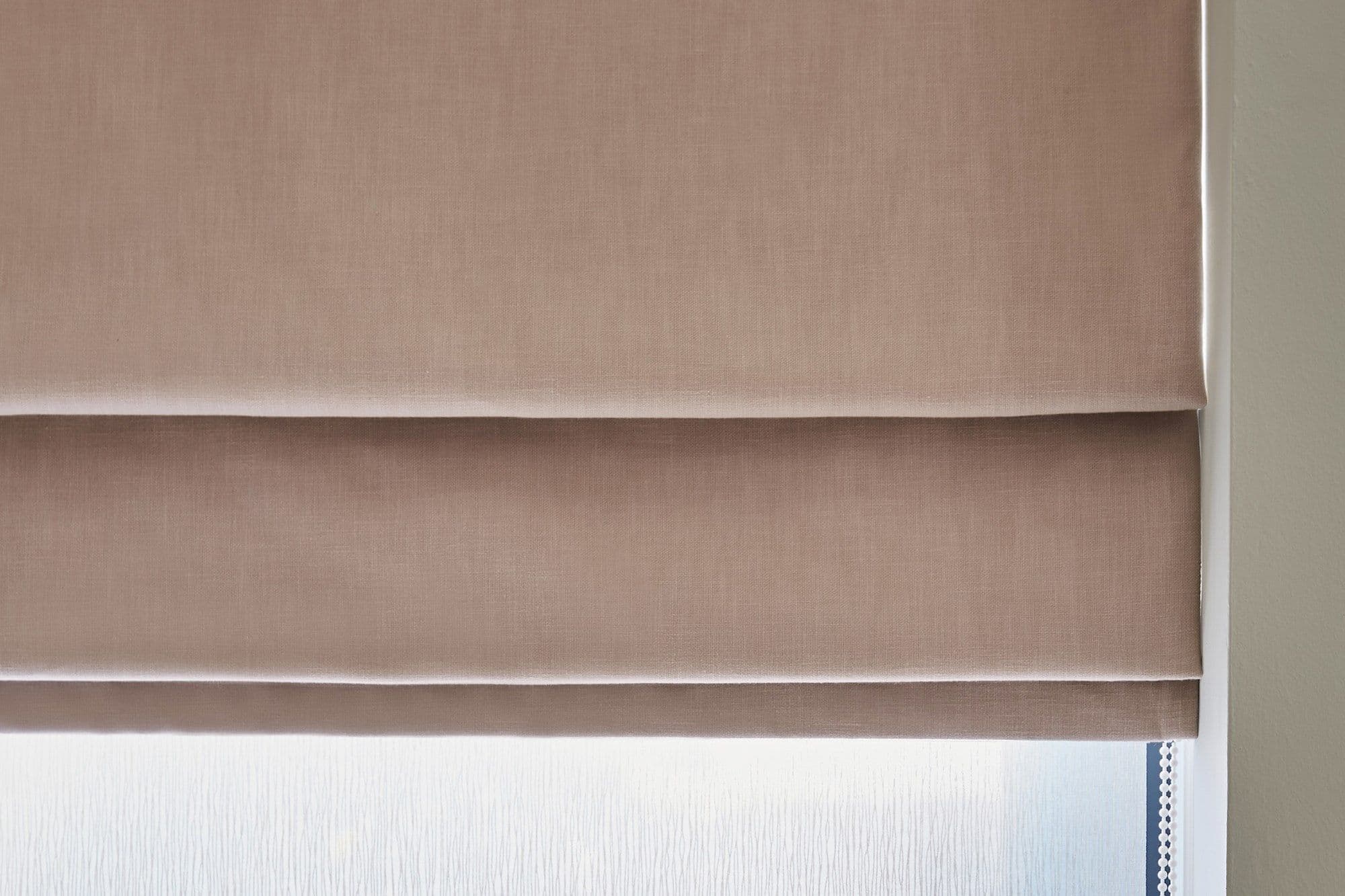 Beige fabric Roman blind, lowered in horizontal tiers, closed and gently filtering daylight, covering a frosted window in a neutral-toned interior; beaded pull chain visible at right.
