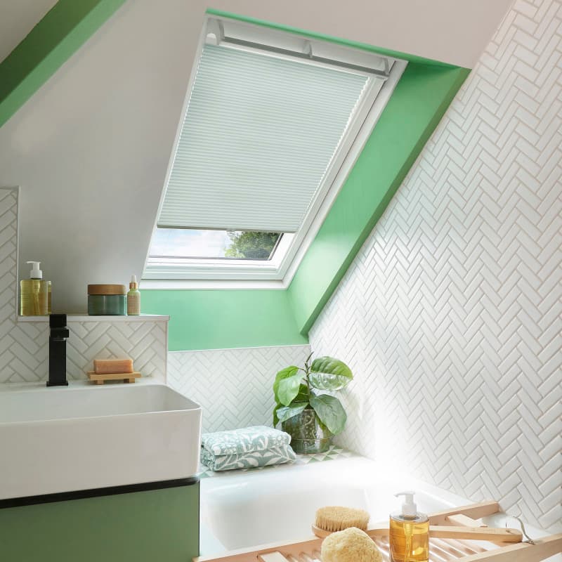 White pleated skylight blind, lowered about two-thirds and filtering daylight; set in a green-painted roof-window reveal above a tiled bathroom with white herringbone walls, sink, bathtub, plant and bath tray.