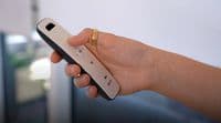 Slim white-and-black remote control held in a right hand, thumb pressing a circular my button; indoor, softly lit room with a blurred window or glass door in the background.

Text: my V somfy
