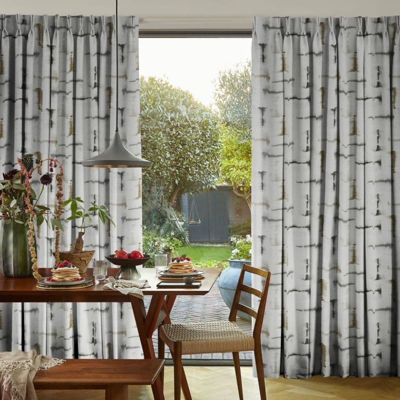 Floor-length pleated curtains with grey birch-print, parted centrally and filtering sunlight, framing a sliding glass door; dining table set for breakfast in a sunlit room opening onto a leafy backyard.