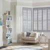 White plantation shutters on a bay window, closed with angled slats filtering sunlight into the room; neutral living room with beige armchair, throw pillows, open shelving and side table.
