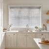 White horizontal Venetian blinds cover a large window, lowered with slats tilted to softly filter daylight; they sit above a farmhouse sink in a bright, neutral kitchen with cream cabinets.