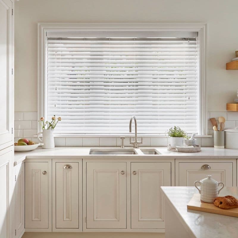 White horizontal Venetian blinds, fully lowered with slats mostly closed, filtering soft daylight into a bright, neutral farmhouse-style kitchen with stainless sink, cream cabinetry, marble counter, plants and utensils.
