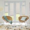 Beige pleated light‑filtering shades lowered over double French doors and narrow flanking windows, softly diffusing daylight; bright modern living room with two woven rattan chairs, pastel throw, and patterned rug.