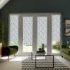 French patio doors fitted with patterned blue-and-white roller blinds, pulled down to mid-height, framing a sunlit garden—modern living room with green armchairs, glass coffee table, chevron rug and plants.