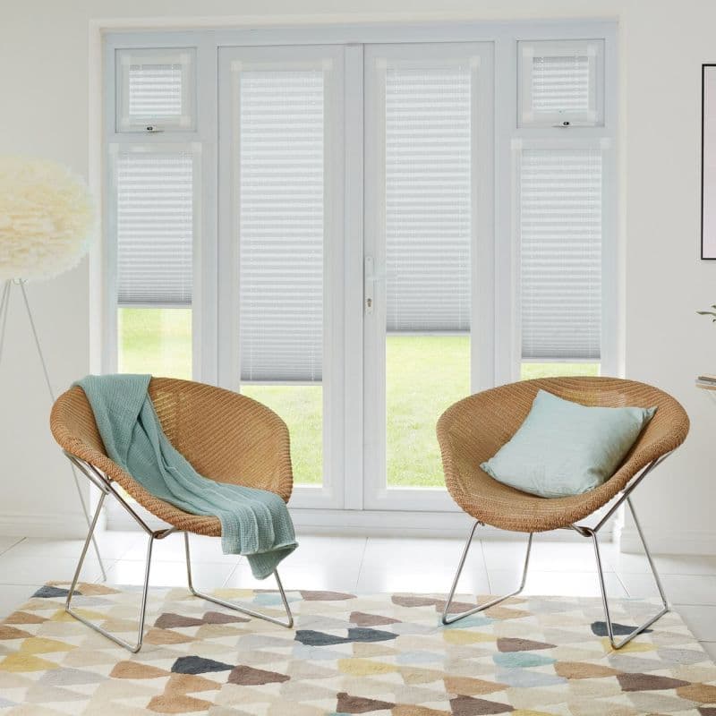 Pleated light-grey blinds on French doors and sidelights, partially lowered and filtering daylight, revealing lawn below; bright living room with two wicker chairs, pastel cushions, geometric rug and floor lamp.