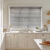 Horizontal Venetian blinds (grey slatted) covering a wide window, closed with slats slightly angled to filter daylight; above a white farmhouse kitchen sink, marble counters, potted herbs and utensils.