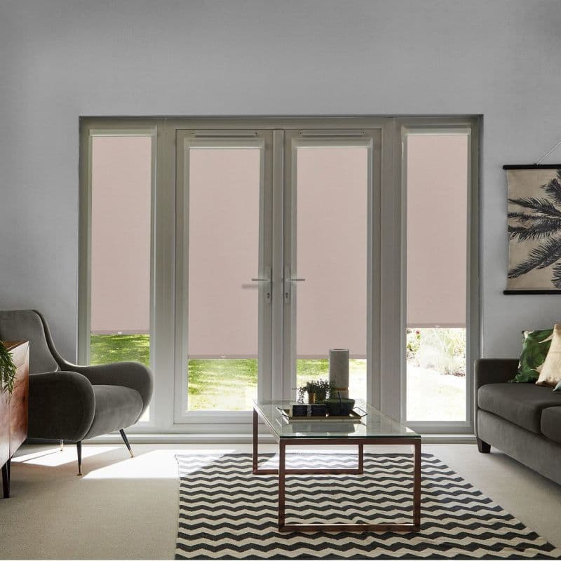 Pink roller blinds cover double French doors and sidelights, lowered to the sill and filtering daylight; living room with grey armchair, sofa, glass coffee table and chevron rug, garden visible.