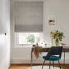 Patterned grey Roman blind covering the window, folded and partially lowered, filtering daylight through a small gap. Context: tidy home office with wooden desk, teal chair, mirror, and vase of flowers.