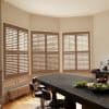 Wooden plantation shutters covering a bay of tall windows, closed with tilted slats filtering soft daylight; overlooking greenery, set above painted wainscoting beside a kitchen island with stools and fruit.