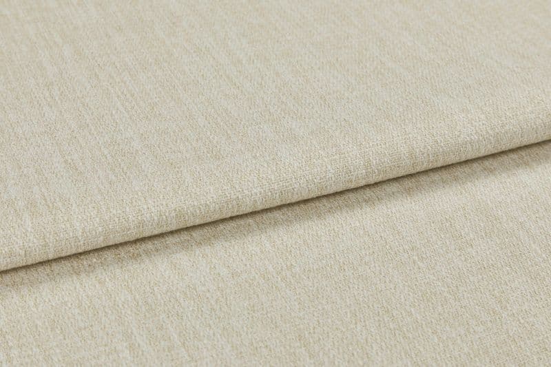 Beige woven upholstery fabric (window dressing absent), folded once with an overlapping edge; close-up shows textured, slightly nubby weave under neutral studio lighting on a flat surface.