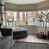 Beige fabric Roman blinds lowered across a three‑panel bay window, softly filtering daylight; decorative vase, candlesticks, potted tree and grey sofa/ottoman in a bright suburban living room.