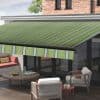 Green striped retractable awning, extended and angled downward, shading glass patio doors and a wooden deck; modern outdoor seating with chairs and low round table with drinks, brick upper wall, daylight.