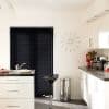 Black horizontal venetian blinds covering a sliding door, fully closed and blocking light — in a bright white modern kitchen with stainless stool, trash bin, coffee machine, and red tulips.