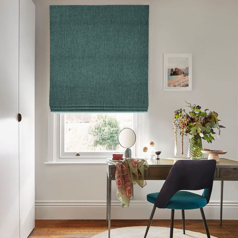 Teal fabric Roman blind lowered over window, filtering daylight; a desk below holds a round mirror, vase of leafy flowers, patterned scarf and teal chair in a softly lit room.
