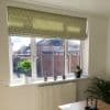 Green leaf-patterned Roman blind, lowered partway and filtering sunlight; three-pane window with sill holding potted plants and candle holders; bright interior overlooking suburban houses and a wooden fence.