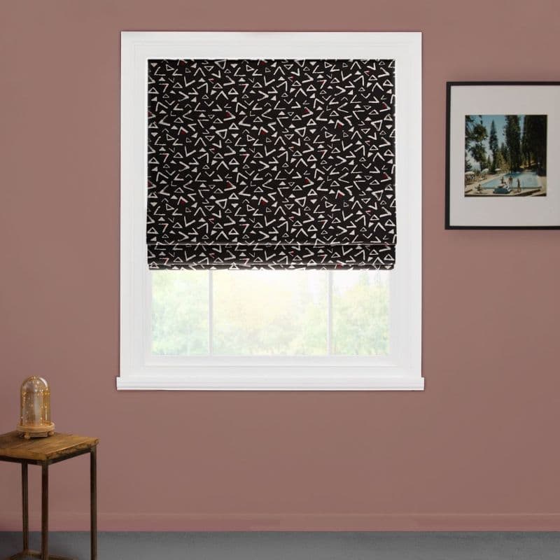Black Roman shade with white triangular pattern and red accents, lowered, filtering daylight; mounted in a white frame on a mauve wall with framed art and a side table.