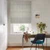 Light-grey patterned Roman blind lowered over window, soft horizontal folds filtering daylight; neutral home office with wooden desk, teal chair, mirror, vase of flowers and framed seaside print.